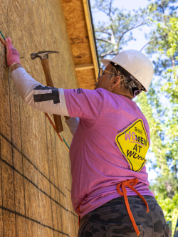 Woman hammers a nail on a Habitat build site while wearing a hard hat and pink shirt that reads "women at work"