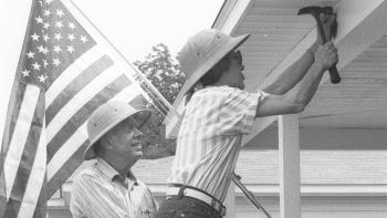 A black and white photo of Mrs. Carter hammering while President Carter looks on. There is an American flag in the background.