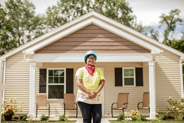 Nusrat, wearing a hard hat and a green "Carter Work Project" t-shirt and nail apron, stands proudly in front of the home she built.