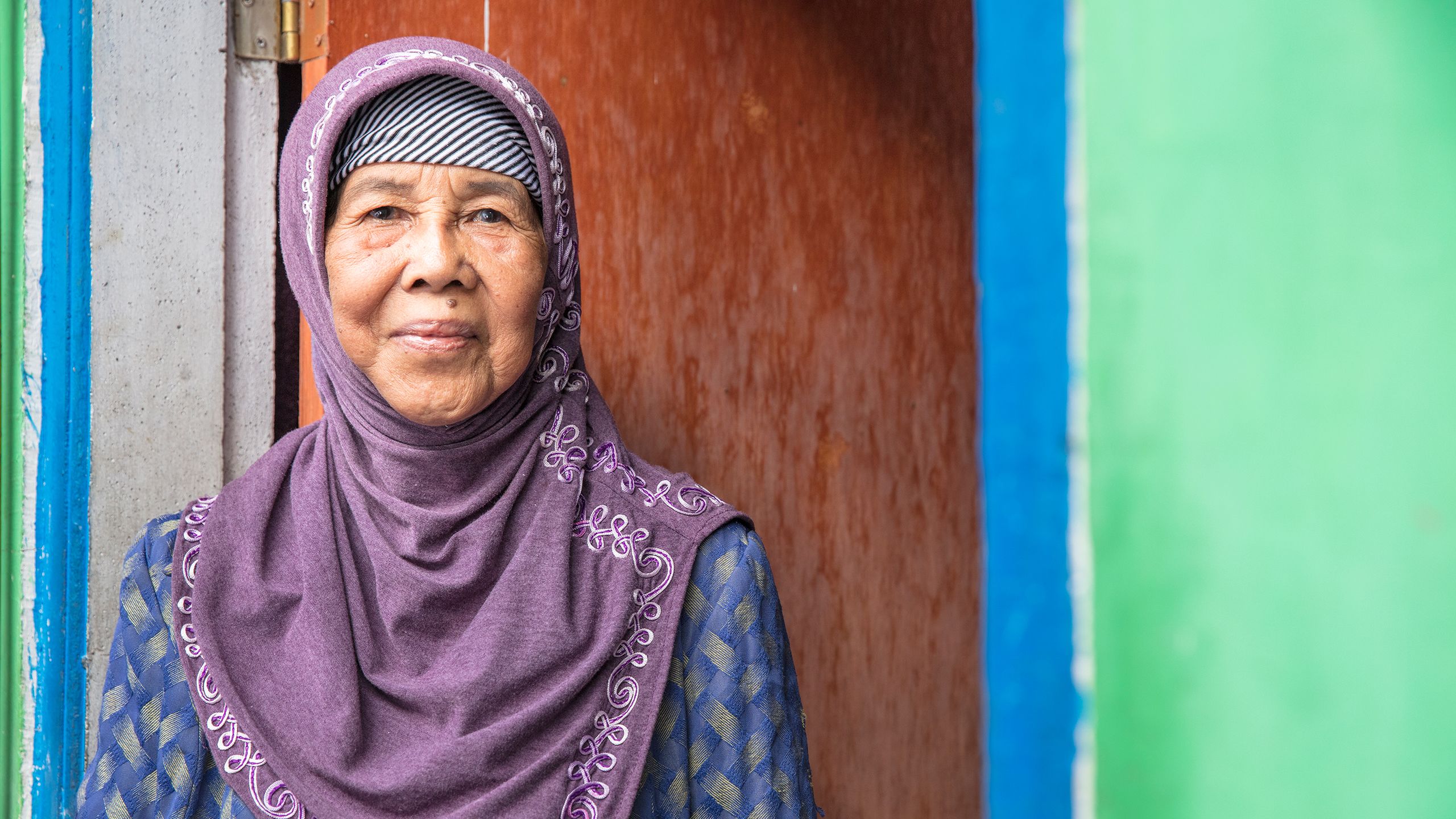 Lusiati, an older woman in a purple headscarf, stands proudly in the doorway of her Habitat house.