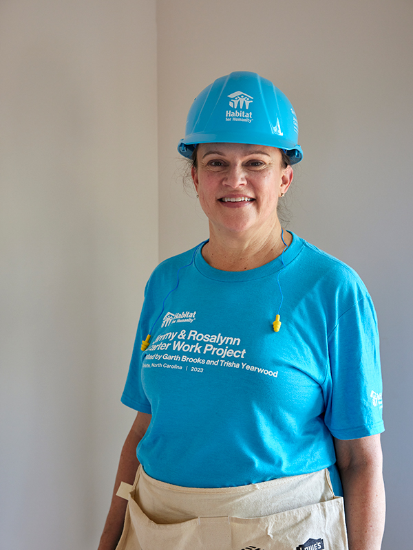 Robin in a blue hardhat and volunteer shirt smiles as she stands inside a Habitat home.