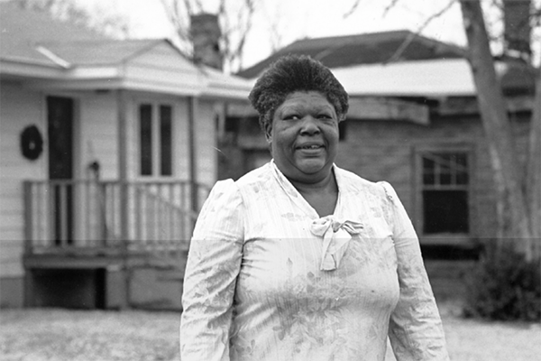 A black and white photo of Delois, a widowed seasonal farmhand, standing outside her home.