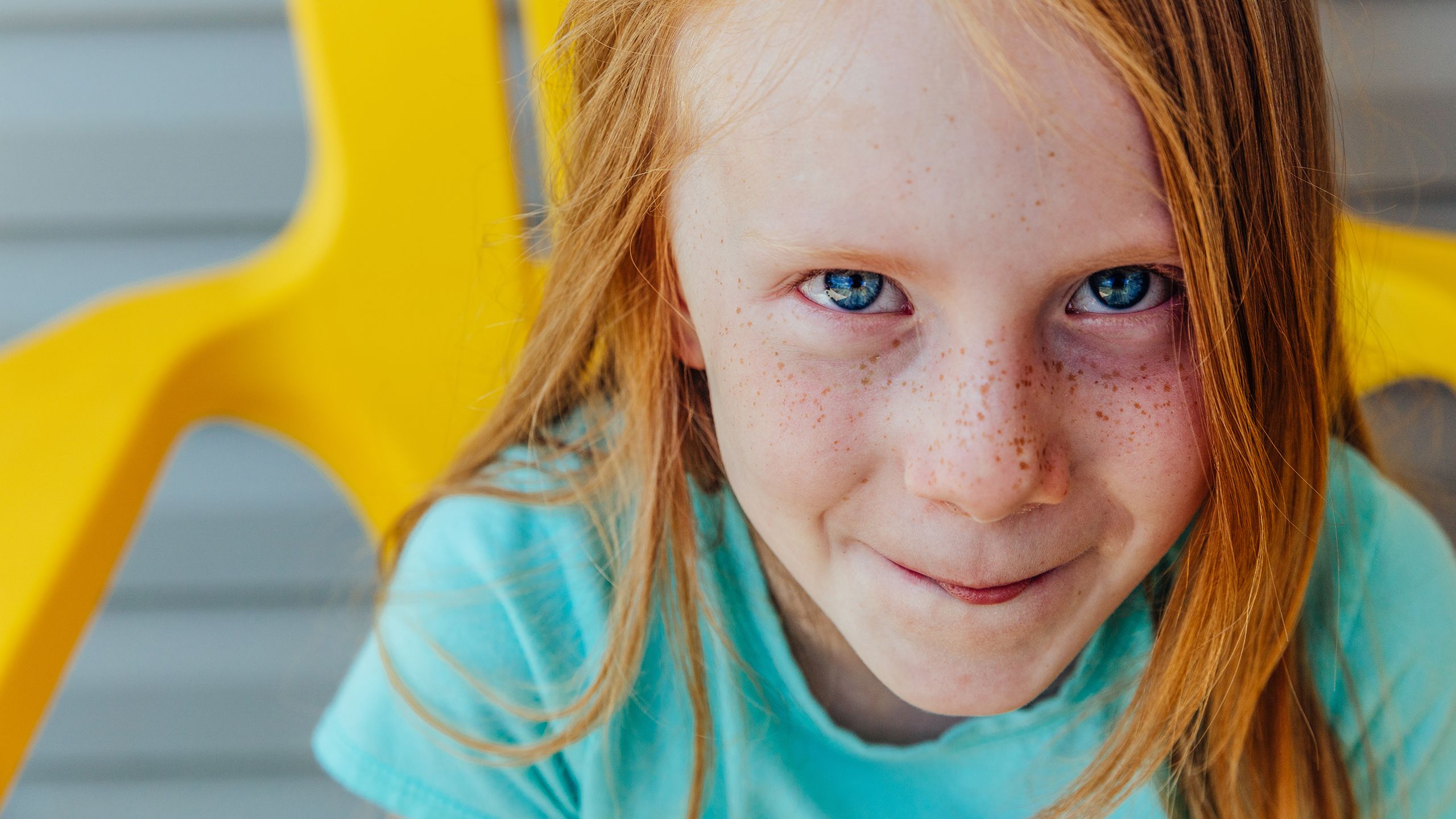 Portrait of Jazmine, a young girl with bright red hair and freckles, in a teal t-shirt sitting in a bright yellow chair.