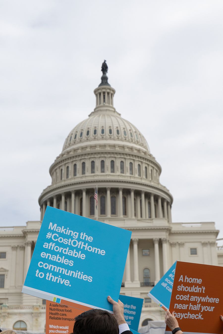 People hold up protest signs in blue and orange in front of the Capitol dome. The front sign reads "Making the #CostOfHome affordable enables communities to thrive."
