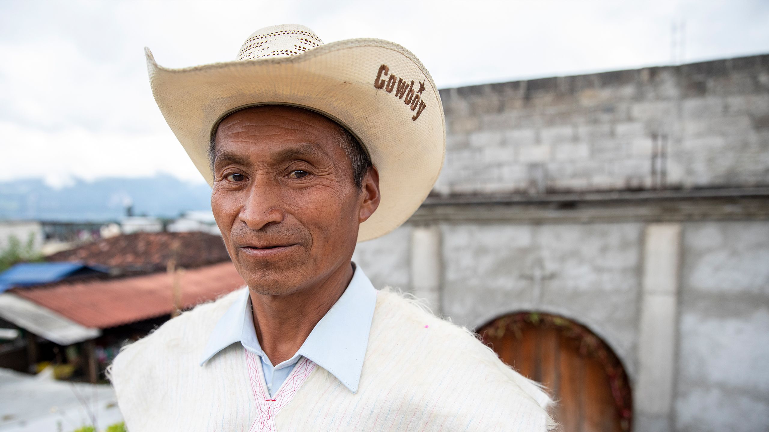 Camilo, a middle-aged Mexican man wearing a straw cowboy hat and wool poncho, stands in front of a cement brick house with the city and mountains in the background.