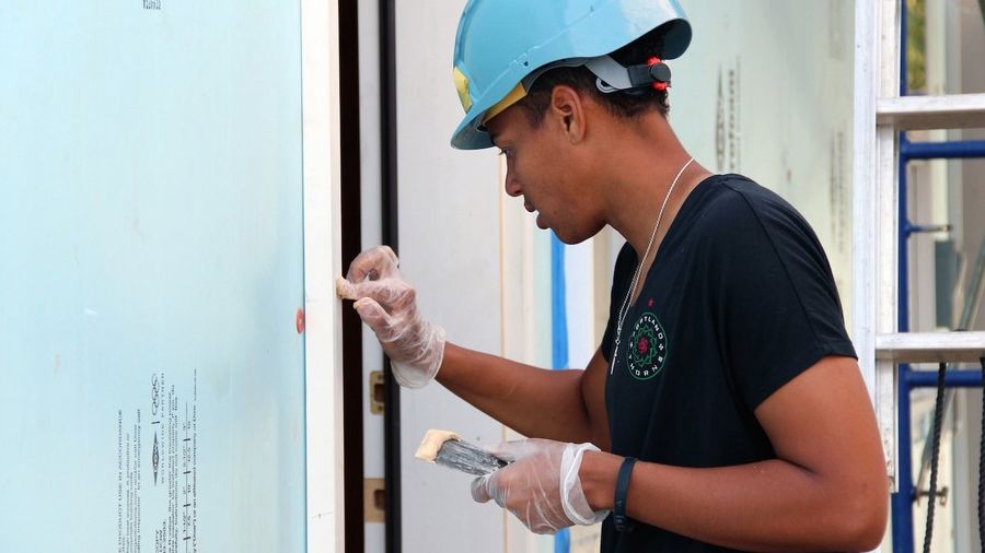 AD, wearing a bright blue hard hat, works to caulk a doorframe on a Habitat house in progress.