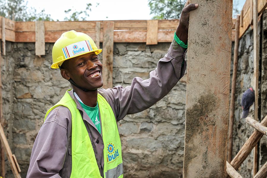 A young man in safety gear with "iBuild" logos holds up a wood plank inside a half-finished stone building.