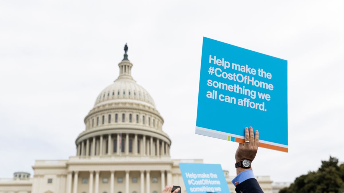In front of the Capitol in Washington D.C., a hand holds a sign reading "Help make the #CostOfHome something we all can afford."