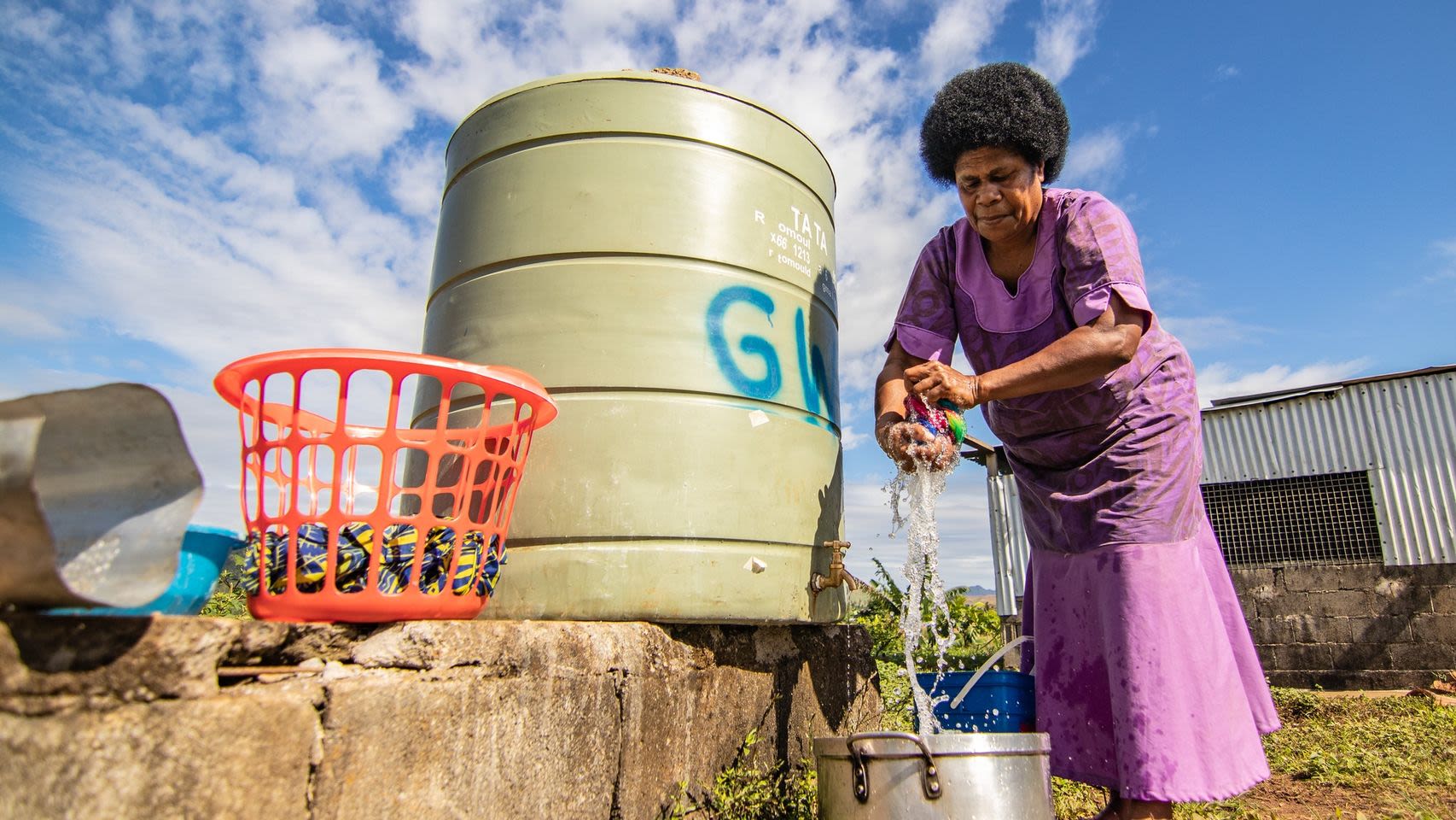 A Fijian woman in a purple dress washes clothing in clean water collected from a rain tank.
