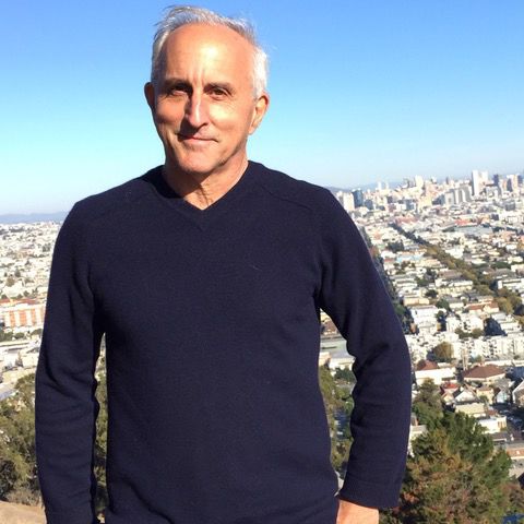 Tony, an older man in a navy sweater, stands outdoors with a view of San Francisco behind him.