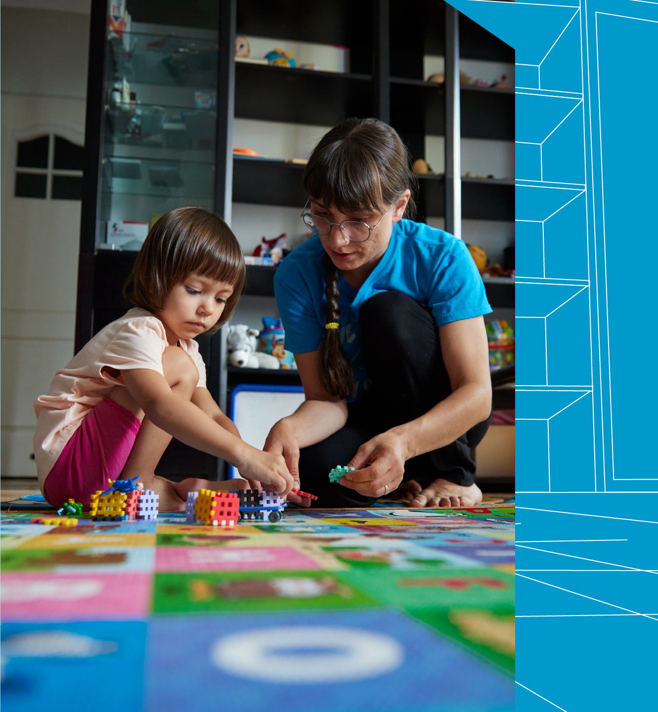 A woman kneels on a colorful play mat with a young girl. The girl stacks a set of building blocks while the woman helps.