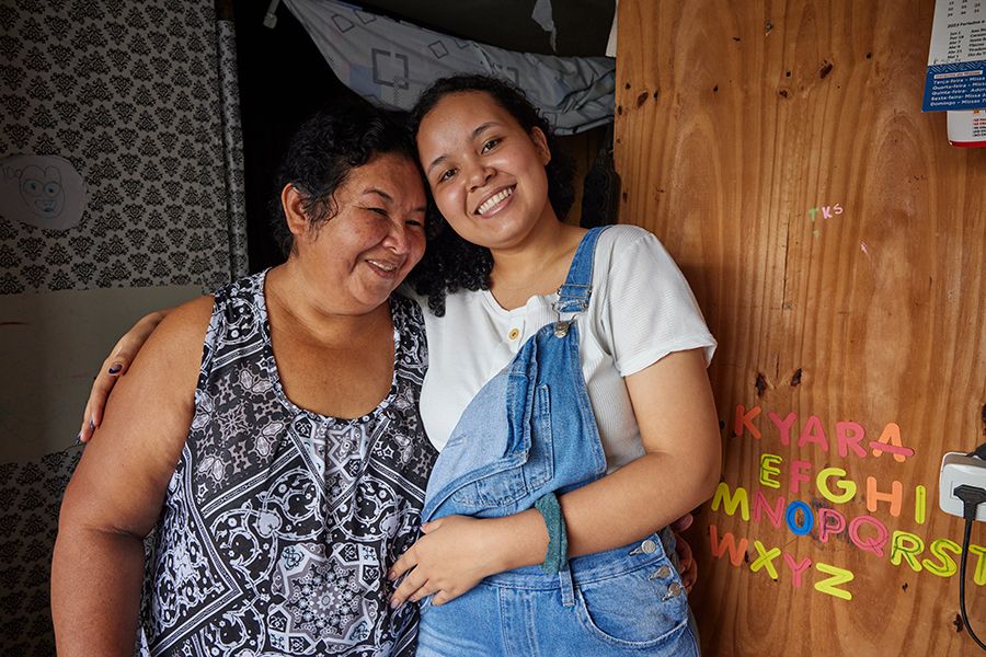 Two Brazilian women hug each other and smile. Next to them is a plywood wall with colorful alphabet stickers.