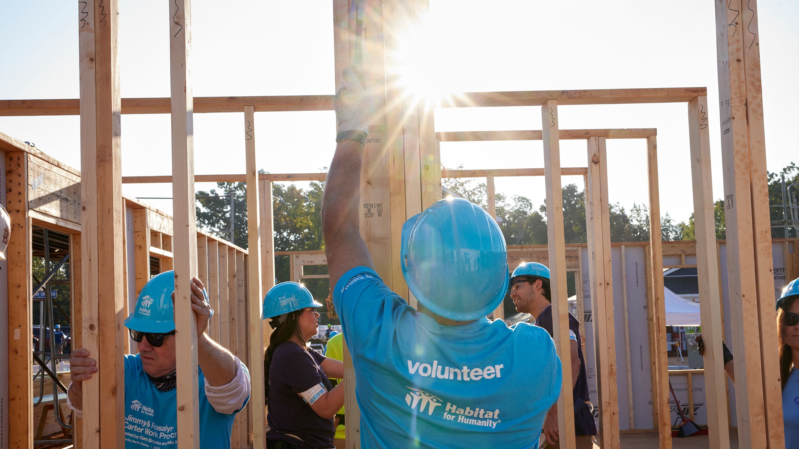 On a busy build site full of volunteers, a man in a hard hat and bright blue t-shirt with "Volunteer" on the back holds a framed wall steady. The sun shines though the open framing in progress.