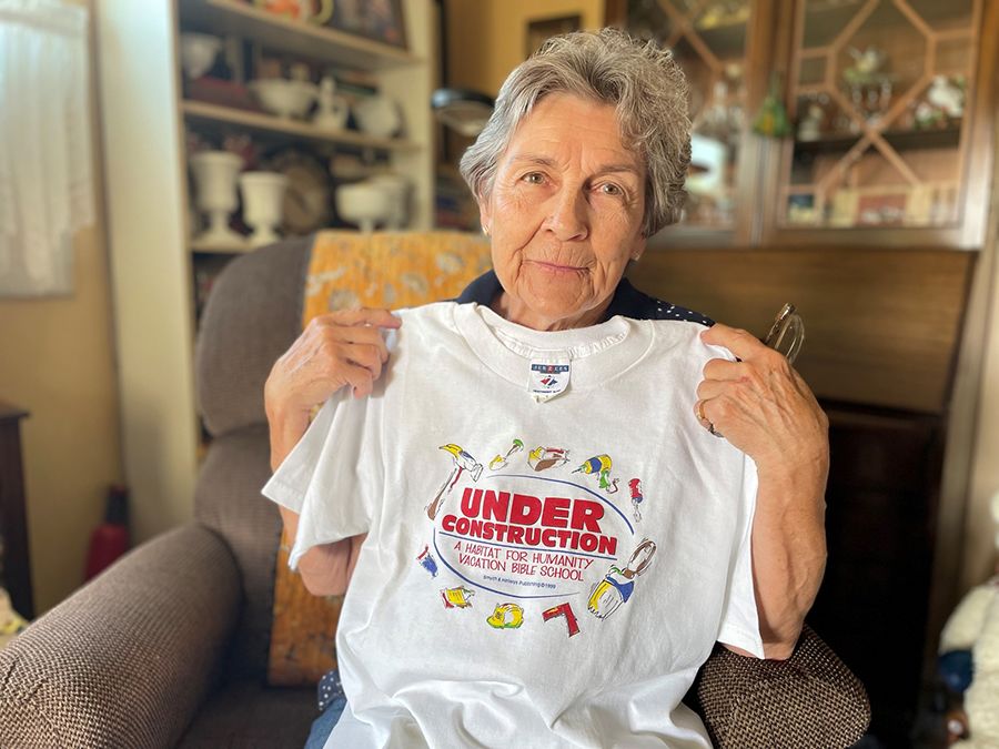 A silver-haired woman sits in an armchair and holds up an old Habitat volunteer t-shirt.