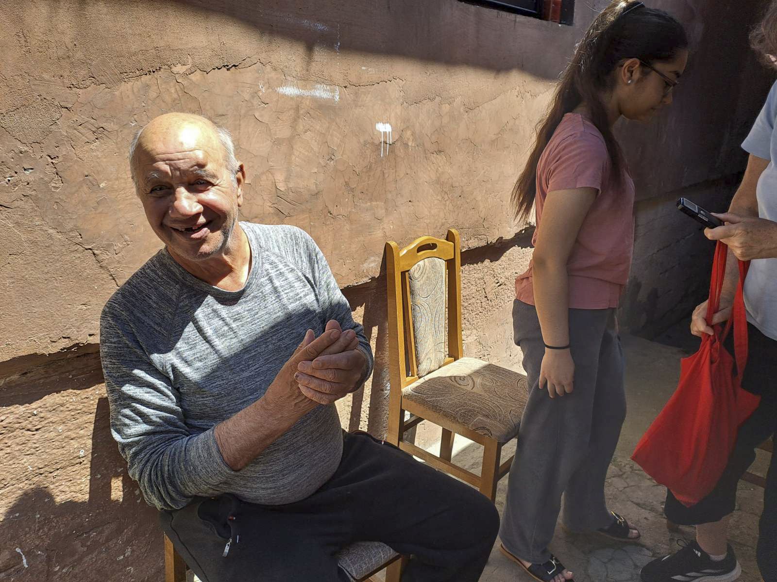 An older Macedonian man sits in a chair next to a plastered wall.