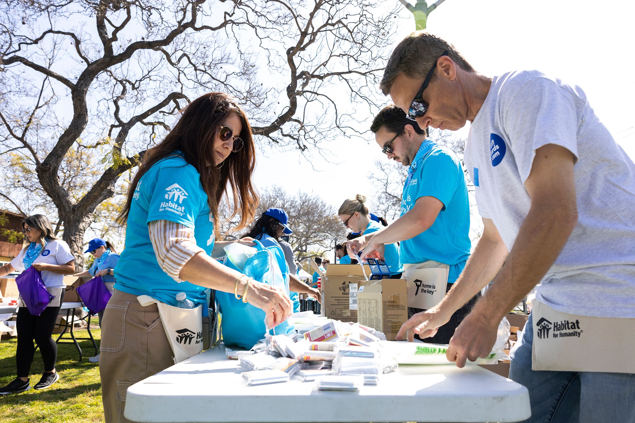 A group of volunteers in bright blue Habitat t-shirts stand along a table outside on a bright day, sorting donated items into bags