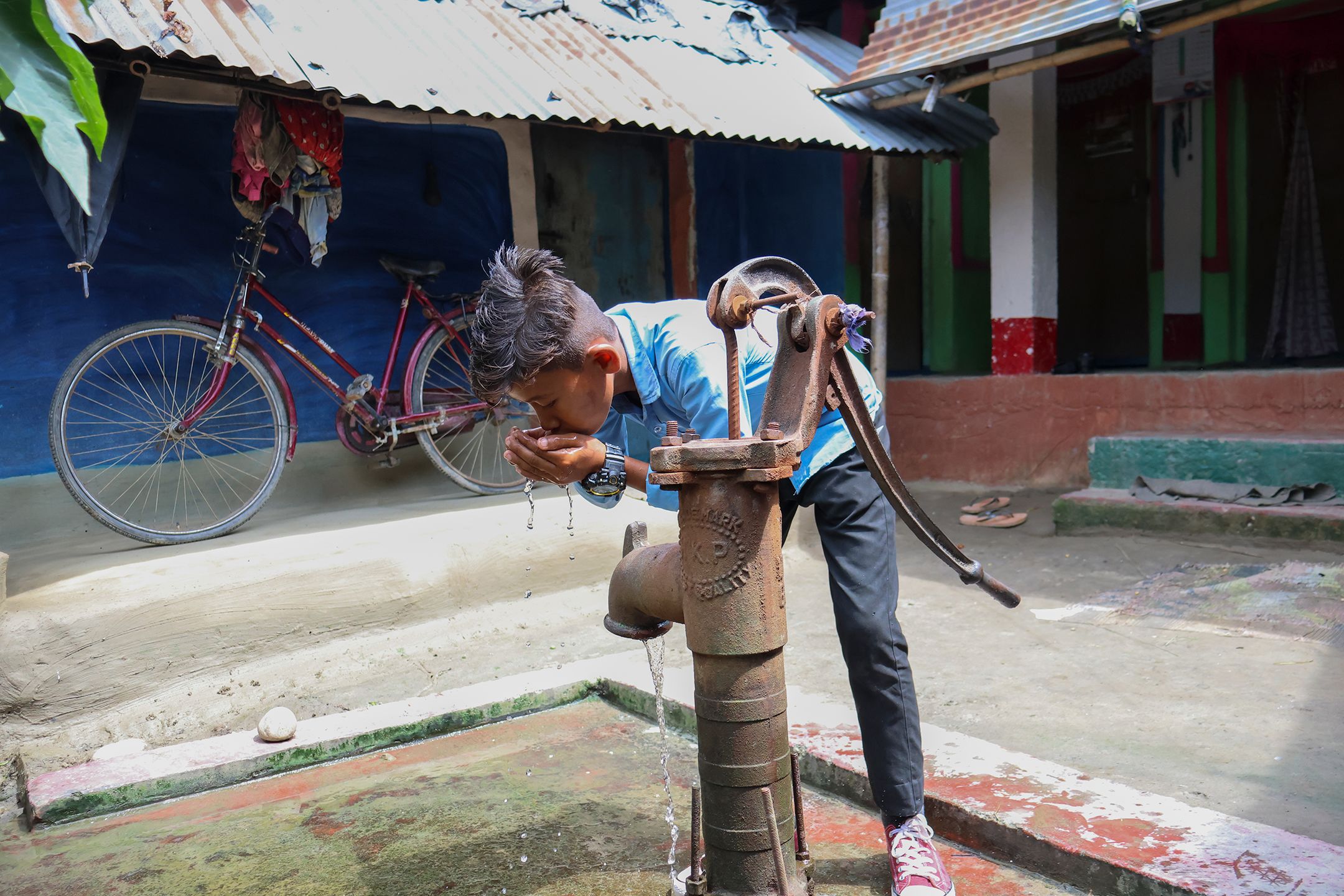 A young Nepali boy drinks water from his cupped hands next to a water pump.