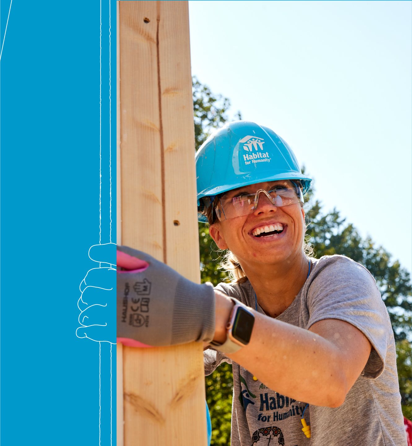 On a sunny build site, a volunteer in a bright blue Habitat hard hat, gray t-shirt and work gloves holds up her end of a framed wall as it's raised. She looks upwards into the sky with a smile.