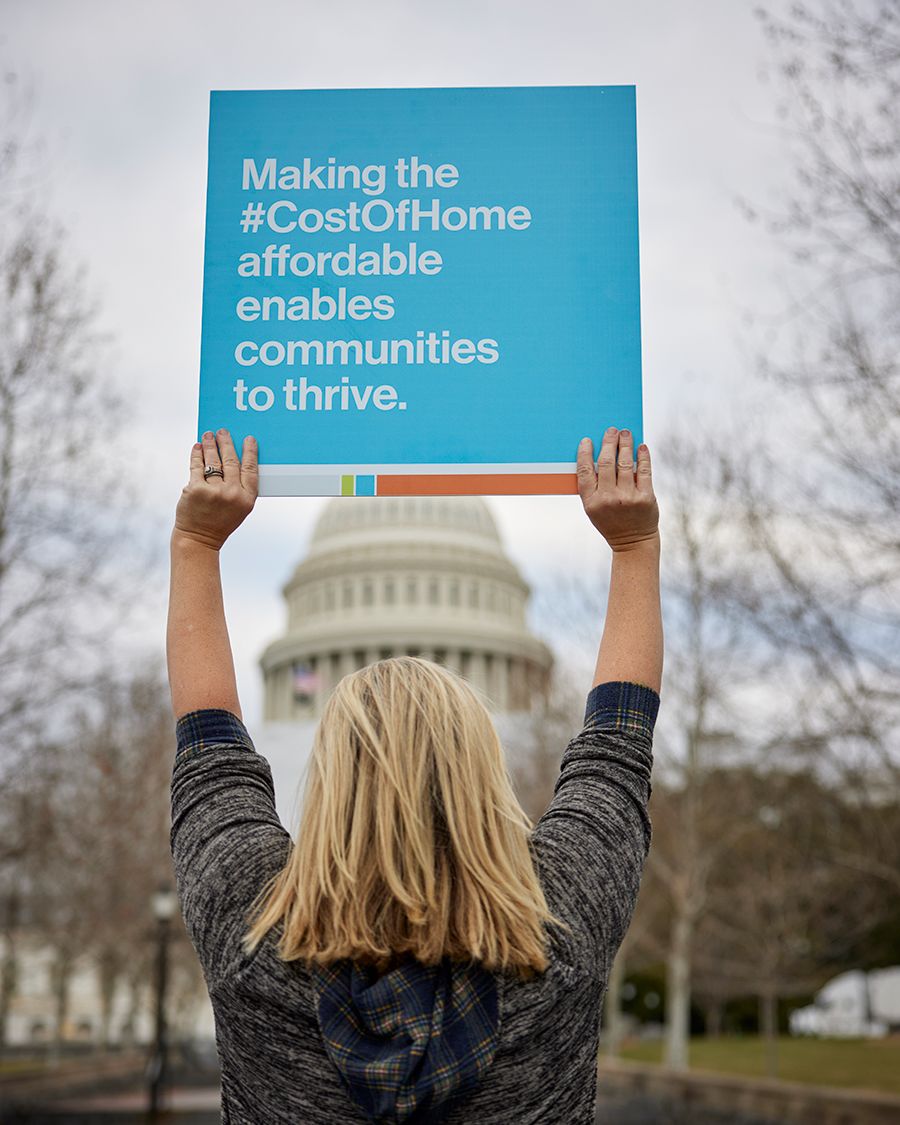 A young woman with blonde hair and a gray sweatshirt holds up a bright blue sign with the Capitol dome in the background. The sign reads: Making the #CostOfHome affordable enables communities to thrive.