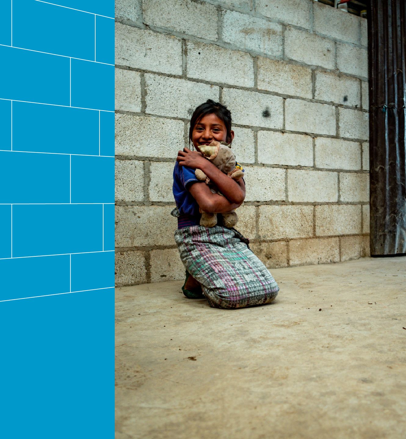 A young girl kneels on a concrete floor. She is smiling and hugging a stuffed animal tightly.