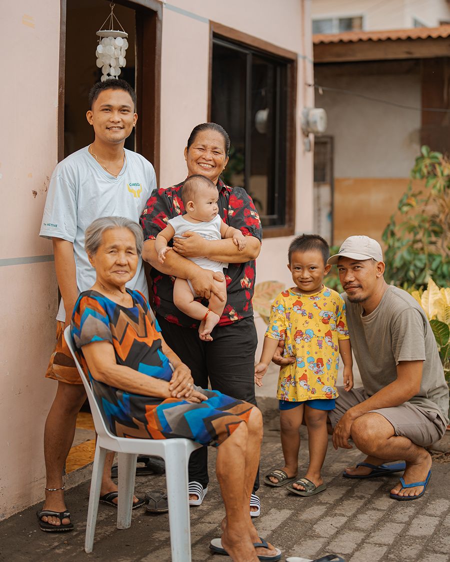 A family sits in front of their home, looking happy and secure. An elderly woman sits in a chair, and behind her are a man and a woman holding an infant. Next to them is another man kneeling beside a young girl.