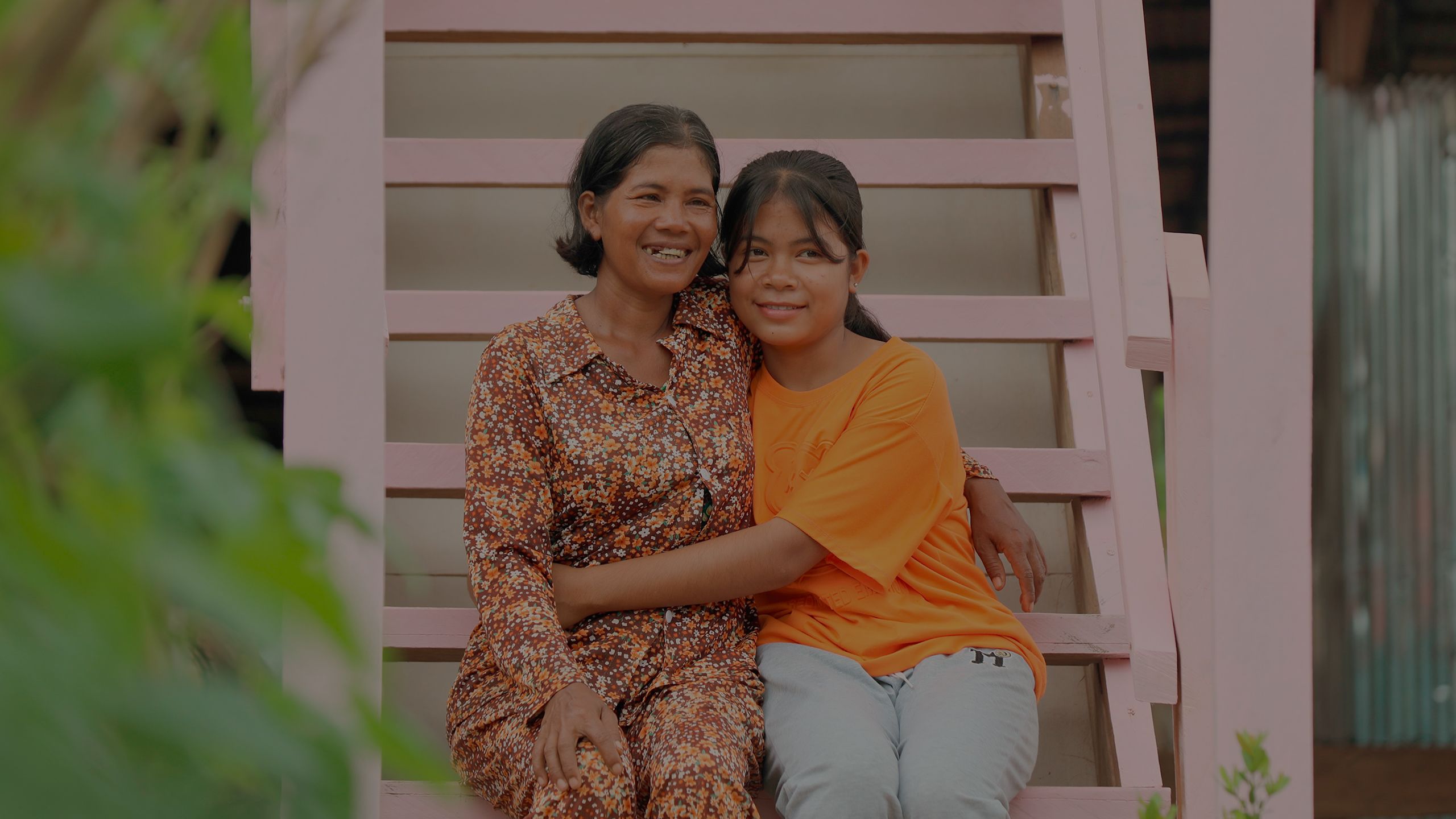 A woman and a young girl hug each other as they sit on the pink stairs of their home.