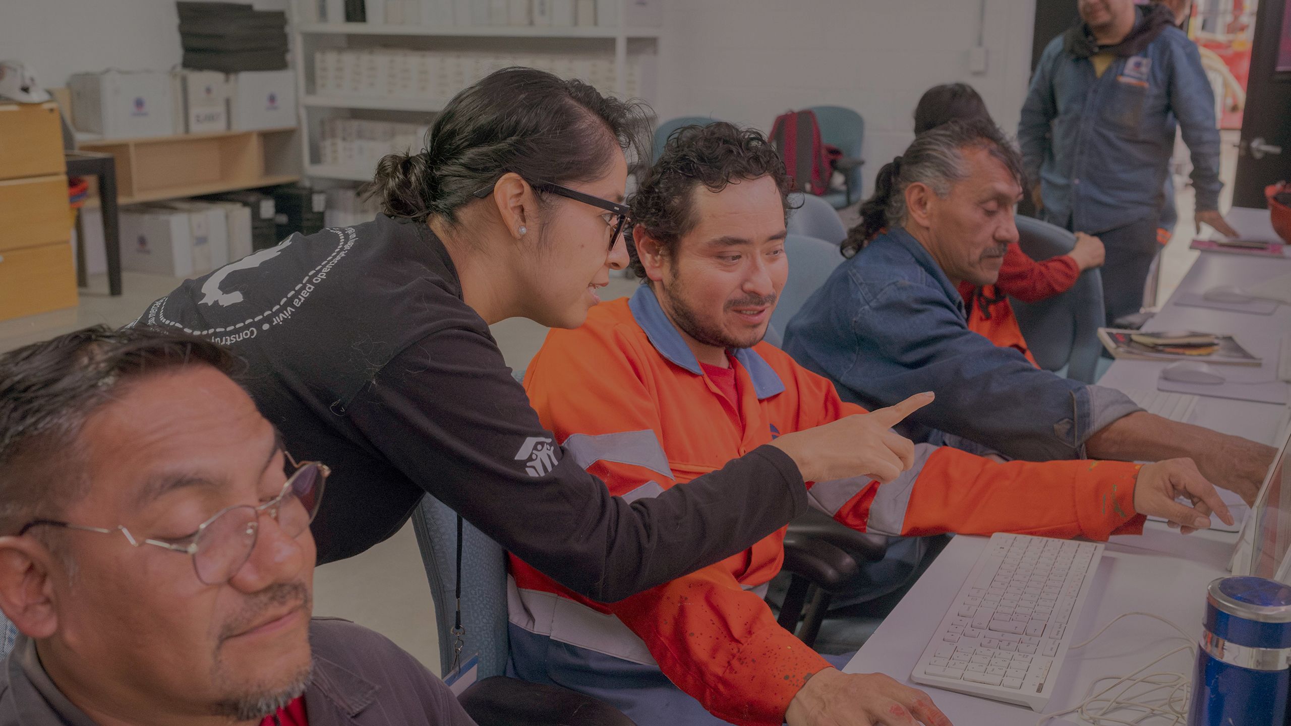 A young man in an orange jacket works on a computer, as a woman in a Habitat t-shirt leans over to point at something on the screen.