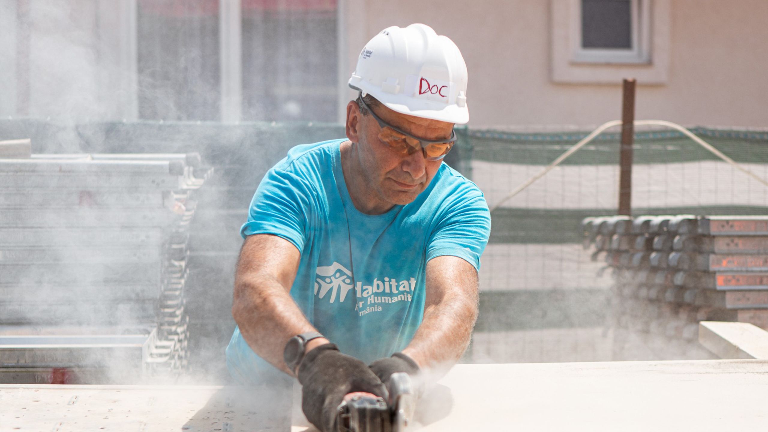 Doc, an older man in a white hard hat with his name on the front, runs a circular saw through plywood on the build site.