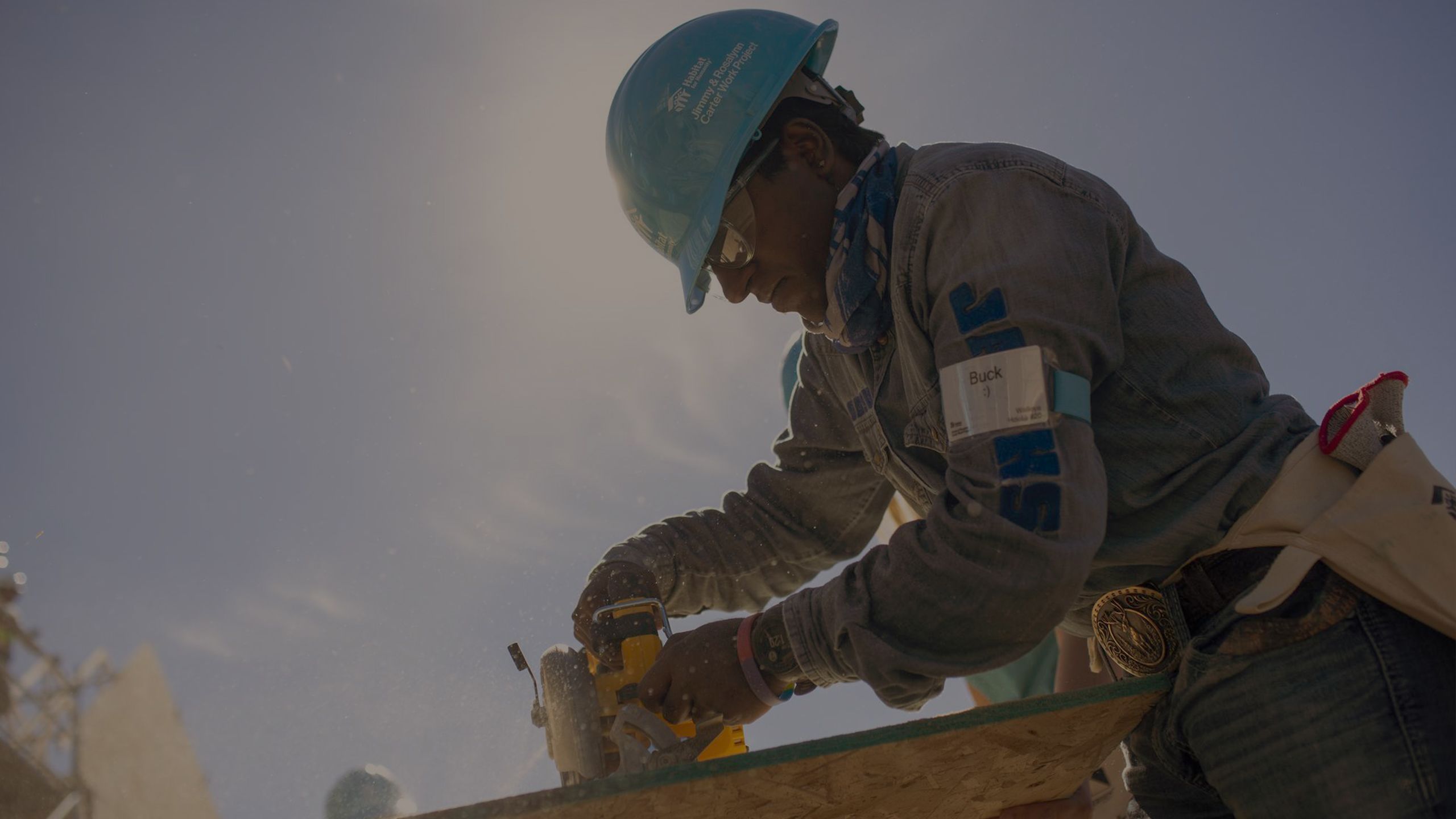 A volunteer in a blue hard hat and long sleeve denim shirt using power tools to cut plywood.