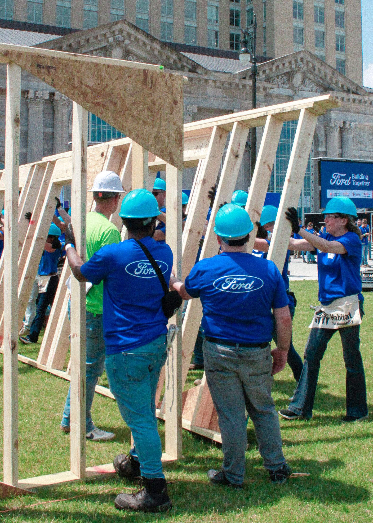 Ford employees standing up the frame of a house with hard hats on. 