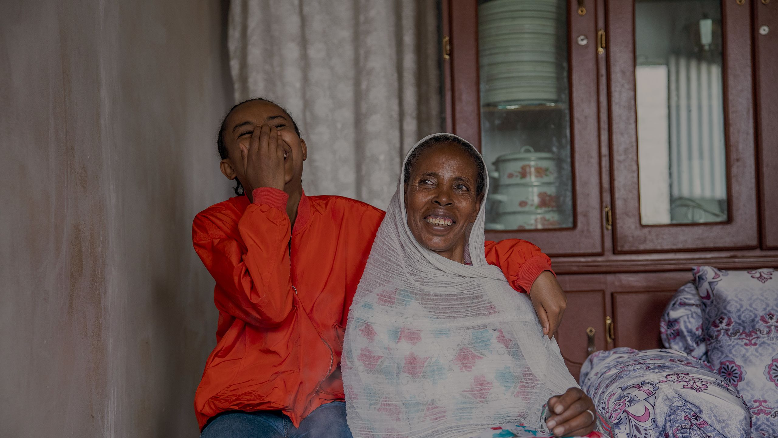 A woman and her daughter sit in their home, smiling and laughing.