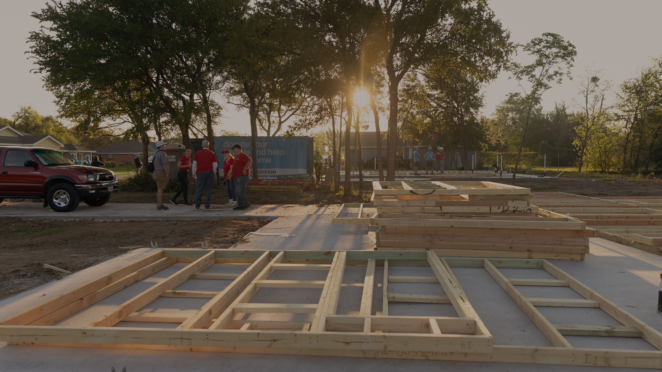 The sun rises over a build site, foundation waiting for walls to be raised. A group of volunteers stand in front of a sign reading: Donate your vehicle to help build a home.