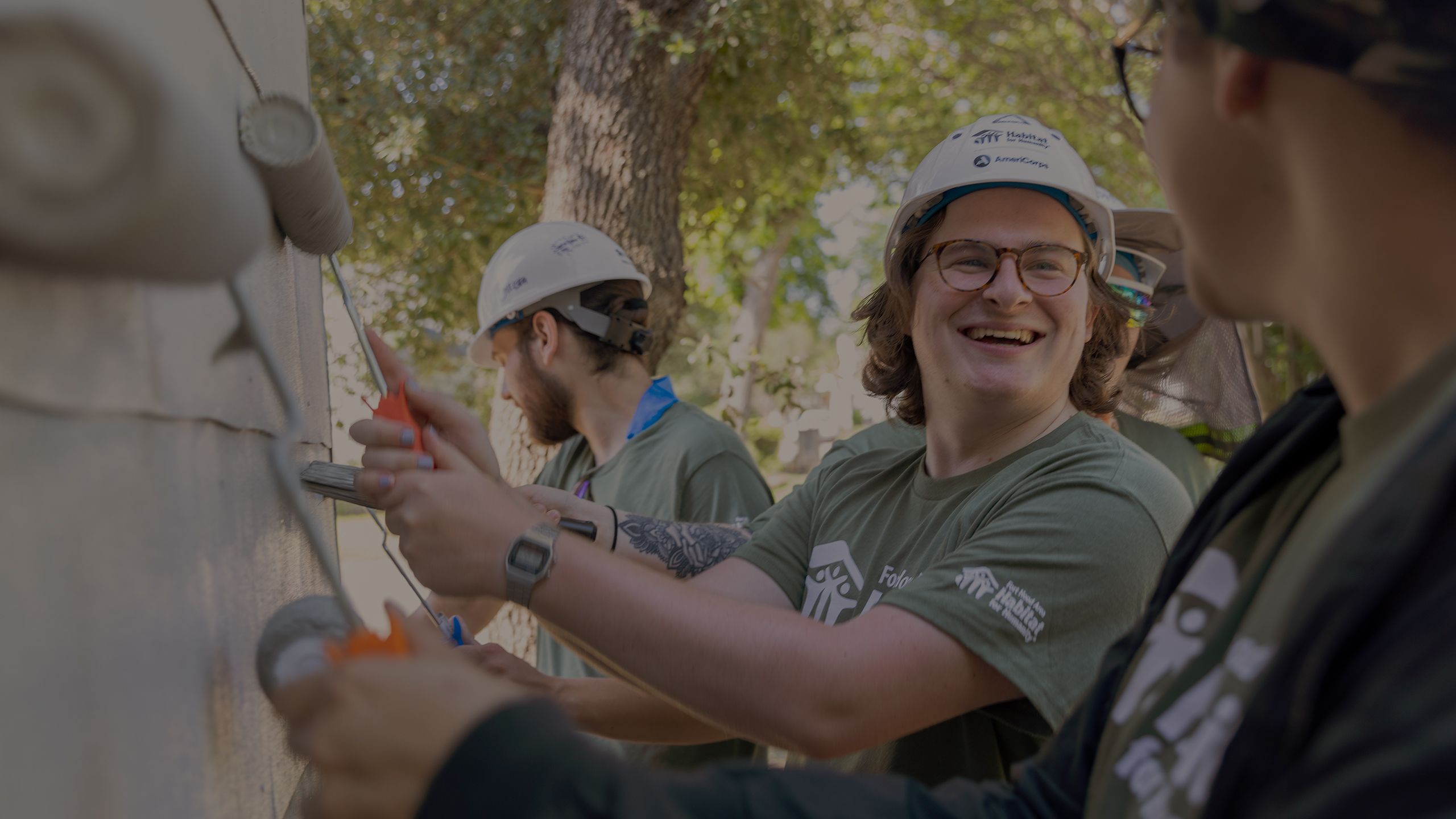 Three volunteers in green t-shirts and white hard hats roll white paint on the side of a home being renovated.