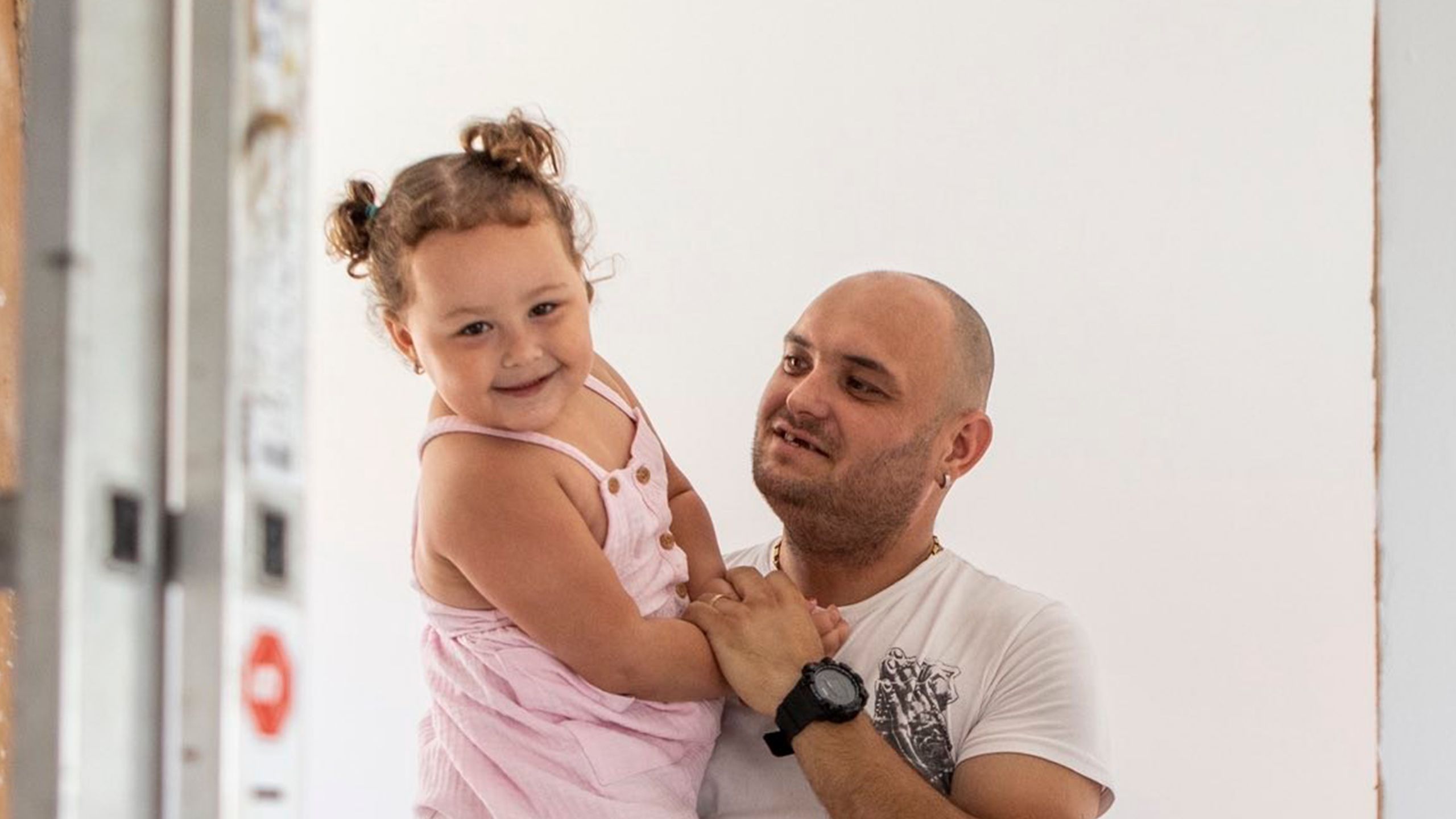 Alexandru, a bald Romanian man in a white t-shirt, holds his young daughter Erika inside their Habitat house. He holds her hand to his heart as she beams at the camera.