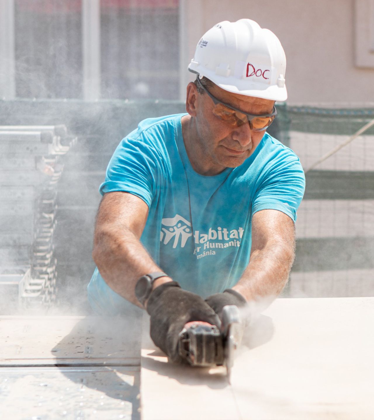 Doc, an older man in a white hard hat and bright blue Habitat Romania shirt, guides a saw through a wallboard.