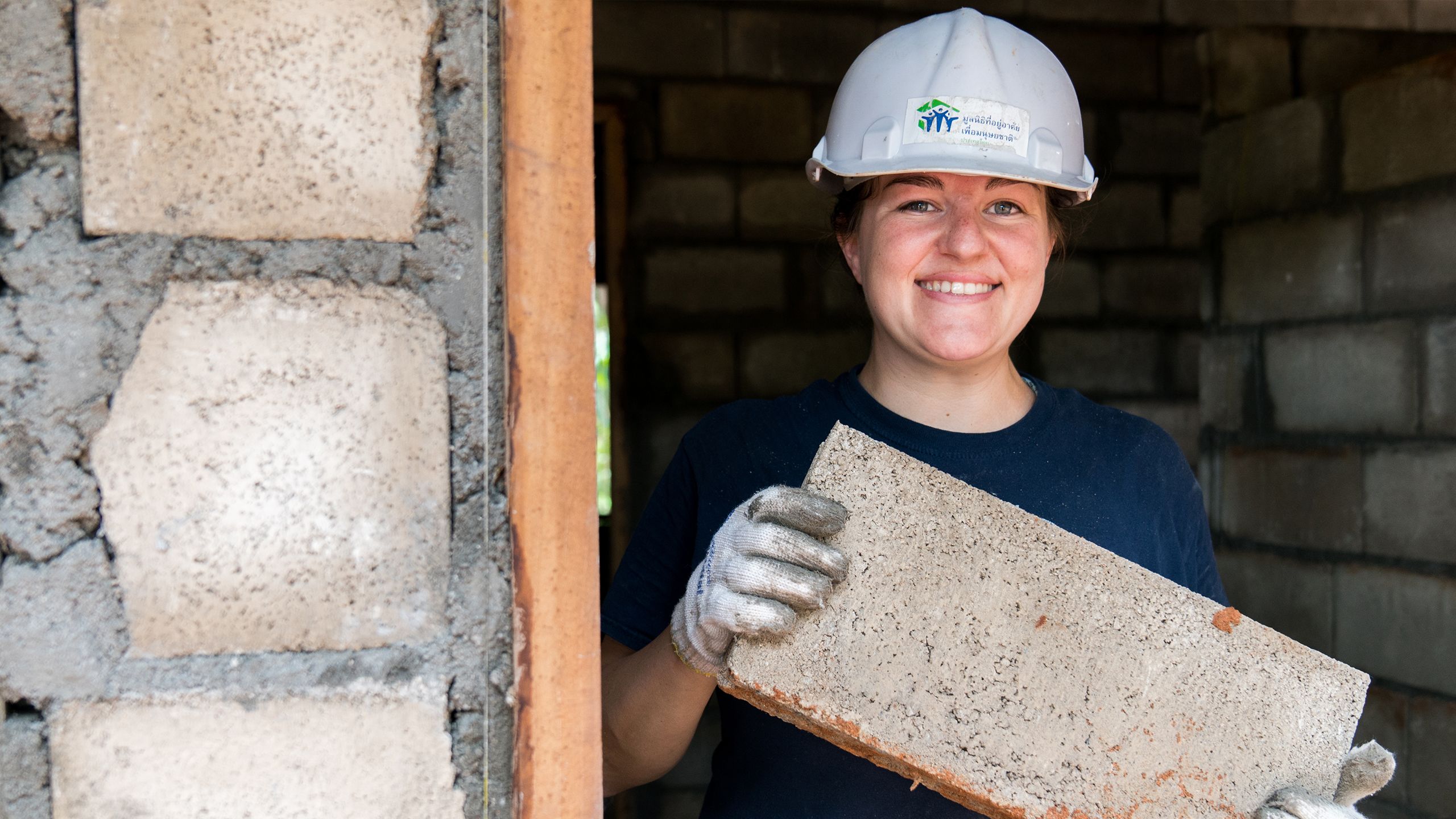 A volunteer in a Habitat hard hat and worn gloves holds a concrete block inside the doorway of a house built with them.