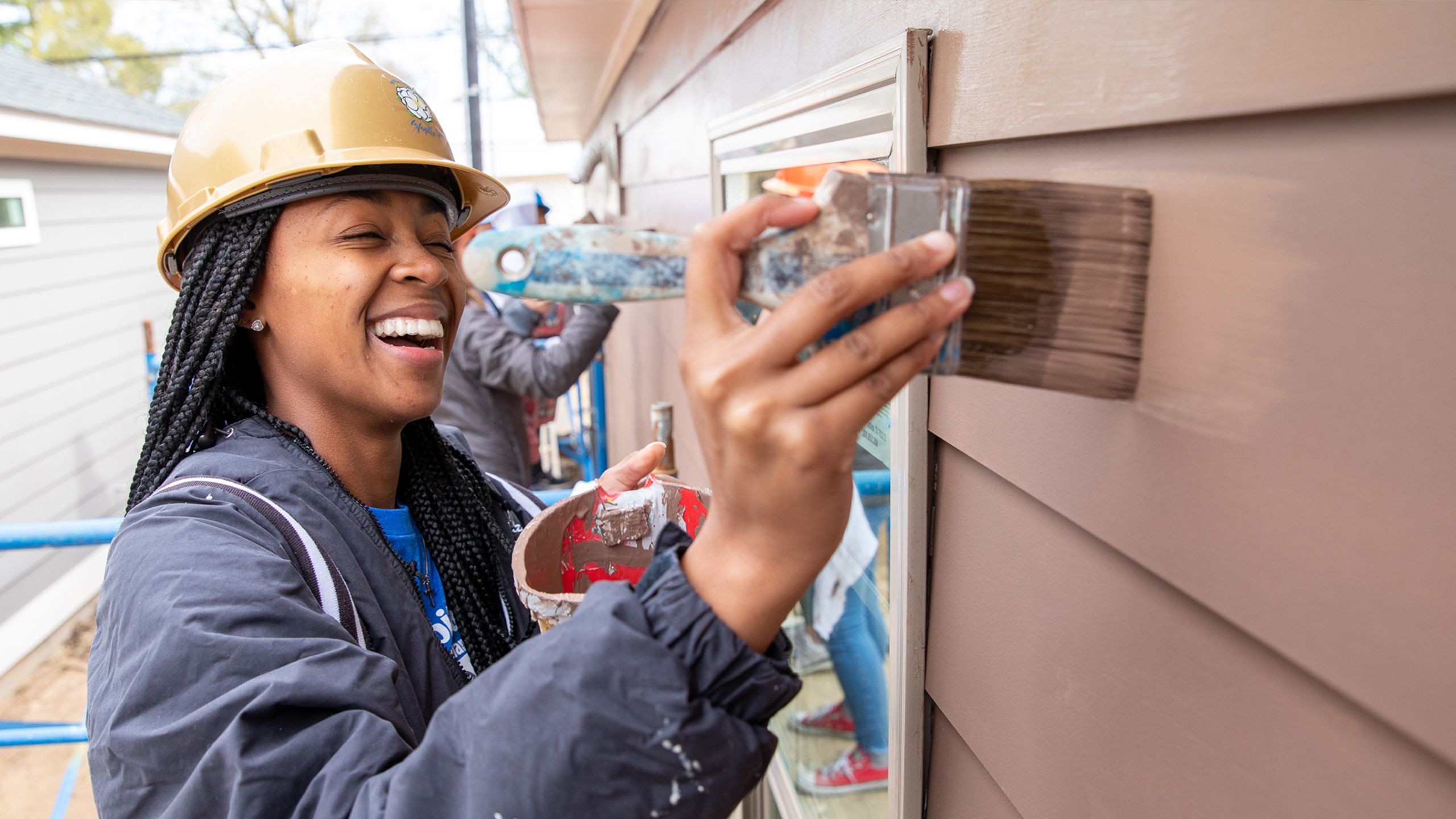 A young Black woman grins while painting the siding of a Habitat home during a Women Build event.