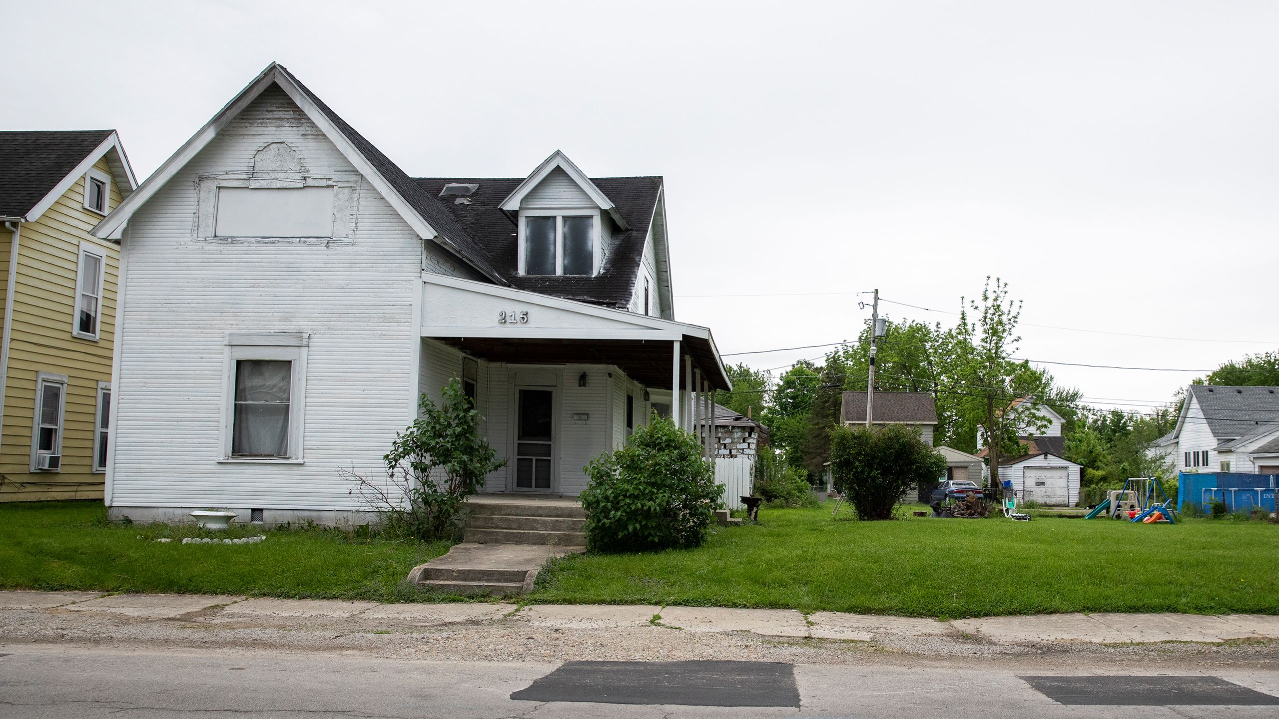 A house with deteriorating siding, poor roofing, boarded-up windows and a struggling yard next to a vacant lot, with crumbling asphalt visible in the road in front.