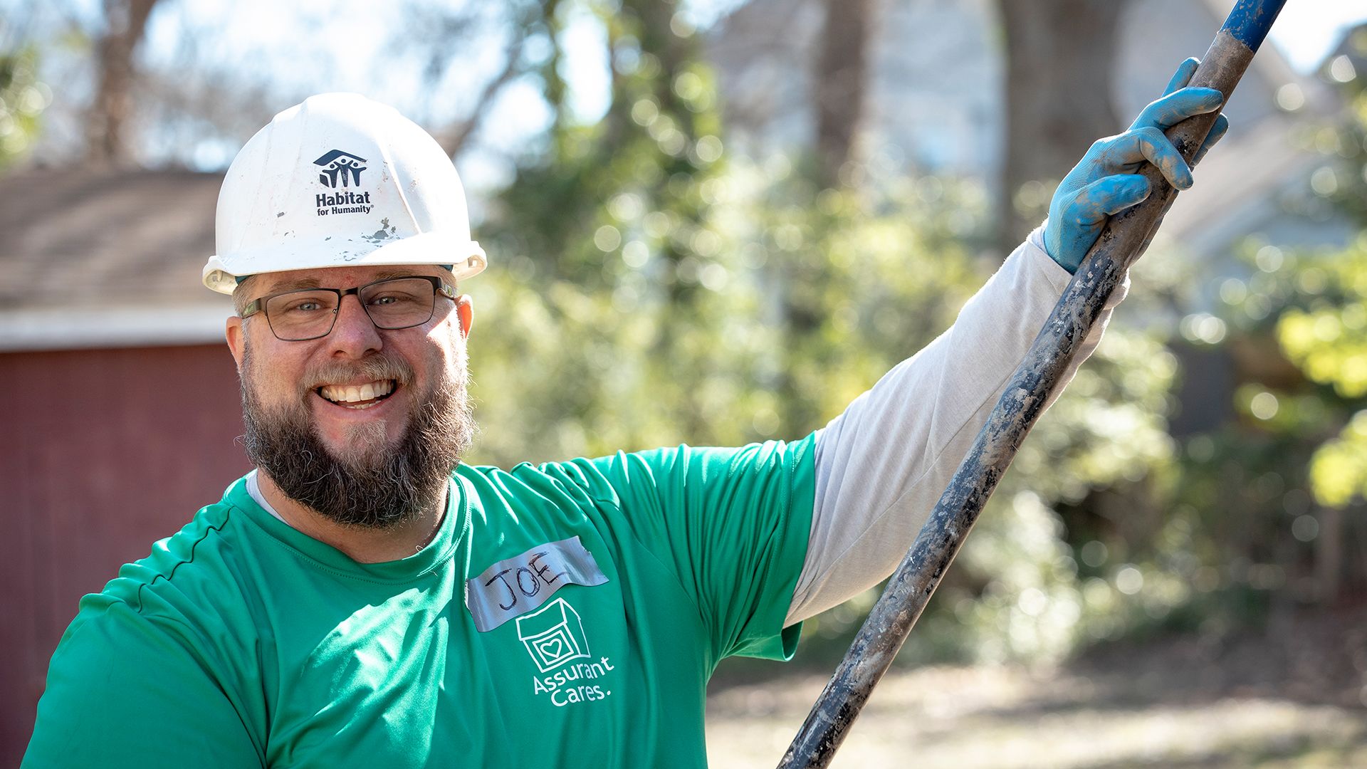 A young man with a beard in a white hard hat and green t-shirt with a duct-tape nametag reading "Joe."