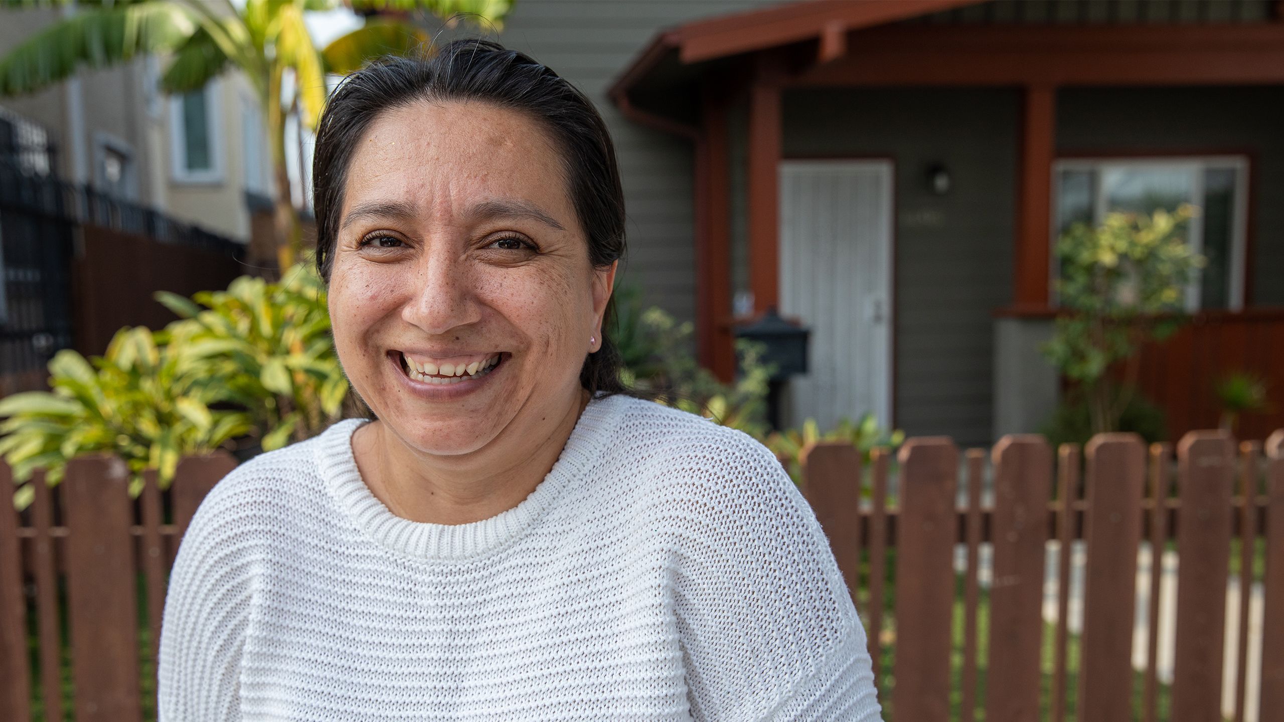 Sonia, a middle-aged woman in a white sweater with olive skin and graying dark hair, beams happily in front of her gray home with banana trees in the yard.