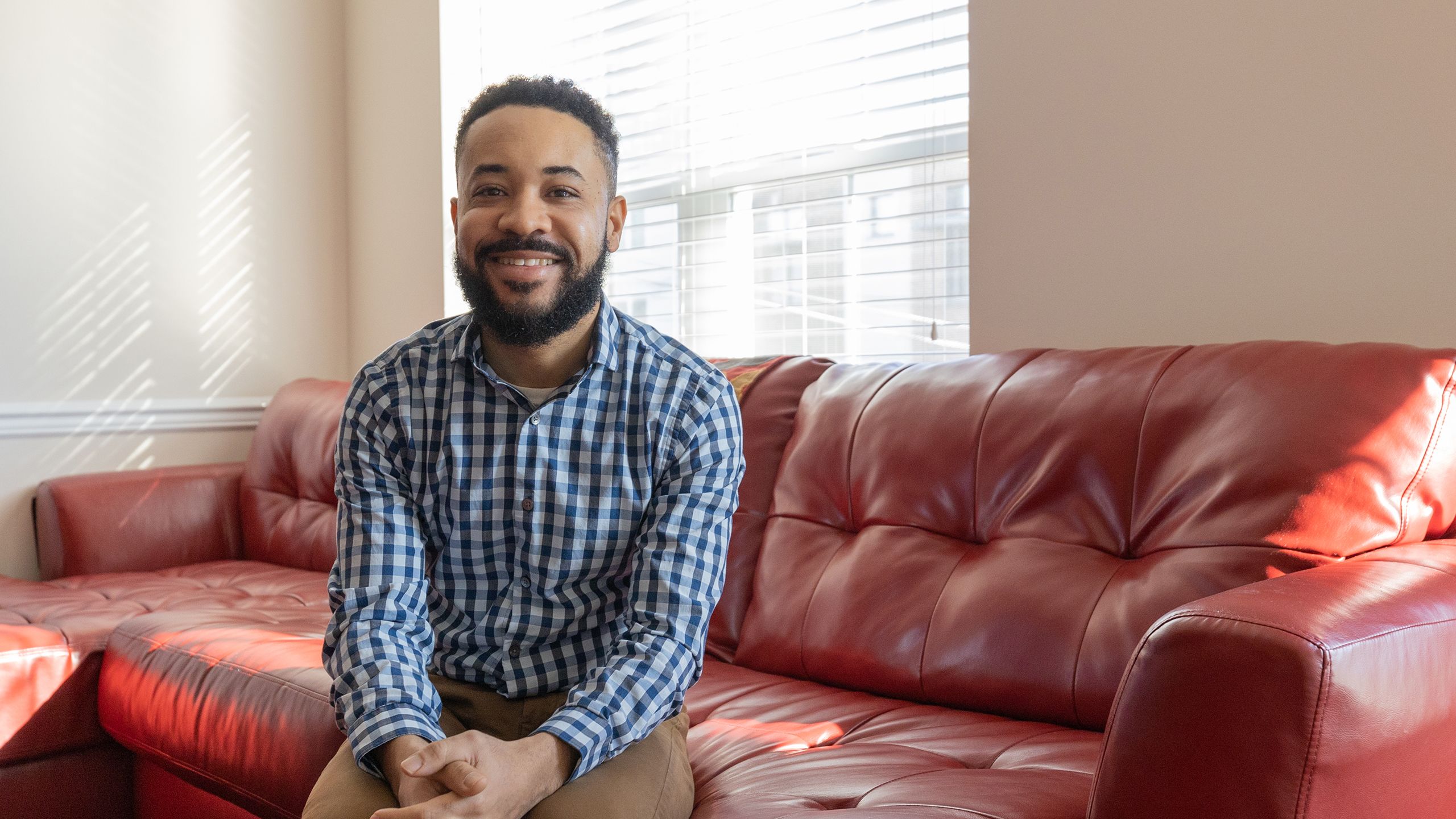 A young man in a plaid button-down sits proudly on the sofa in the living room of his D.C. townhouse in a Habitat-built development.