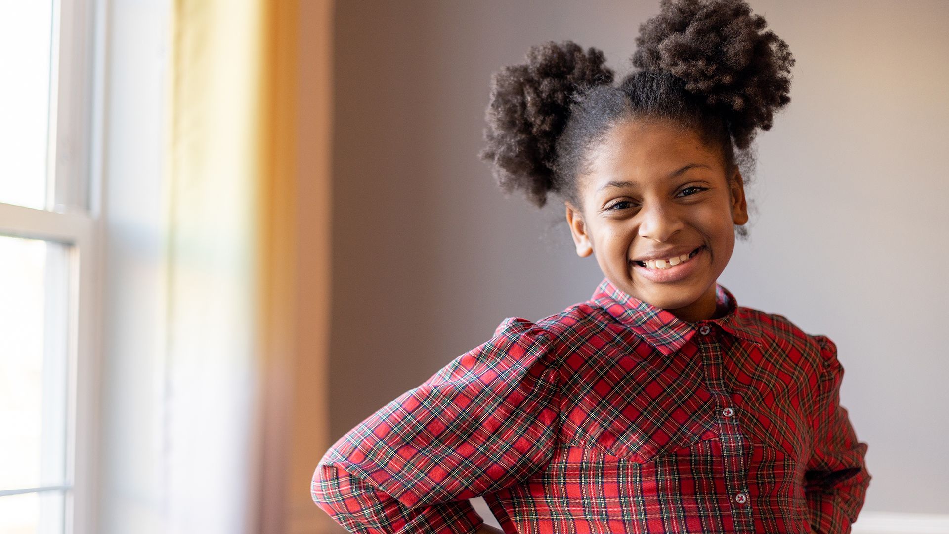 Miracle, a young Black girl with two ponytail puffs and a red plaid shirt, smiles in front of a sunny window.