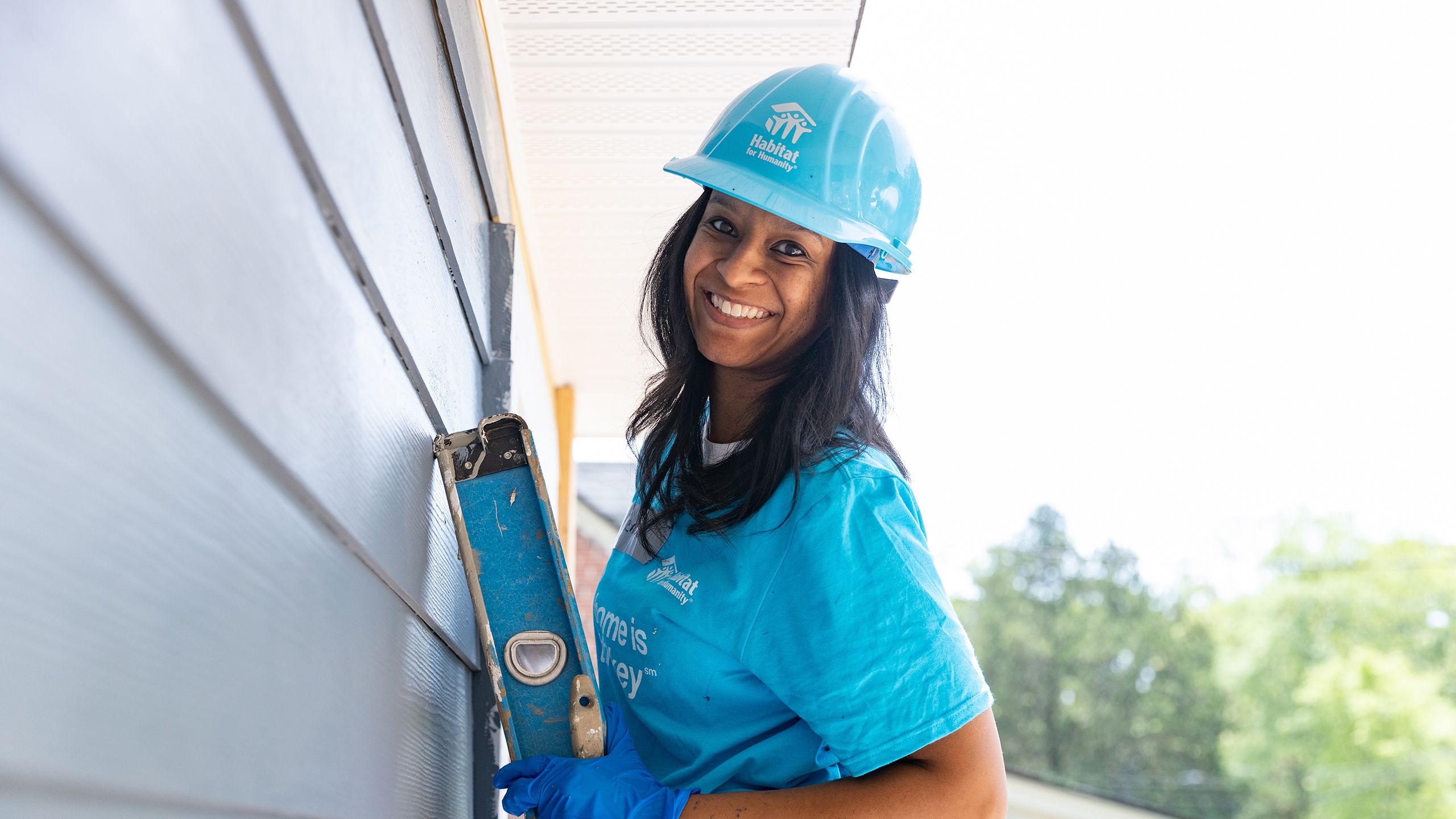 A volunteer in a blue Habitat hard hat smiles and stands on a ladder leaning on a house under construction.