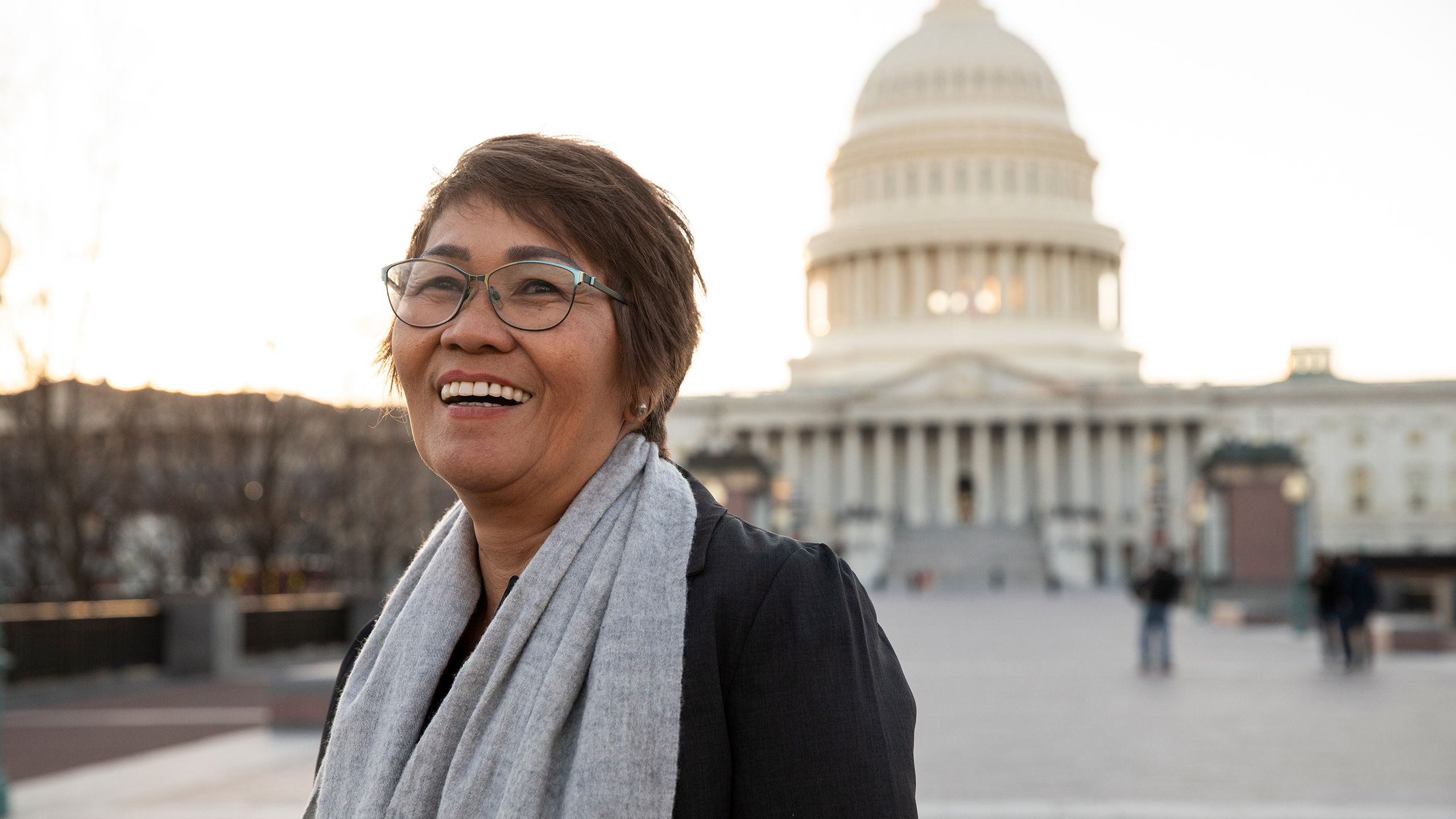 A woman in glasses and a scarf stands in a plaza with the U.S. Capitol building in the background.