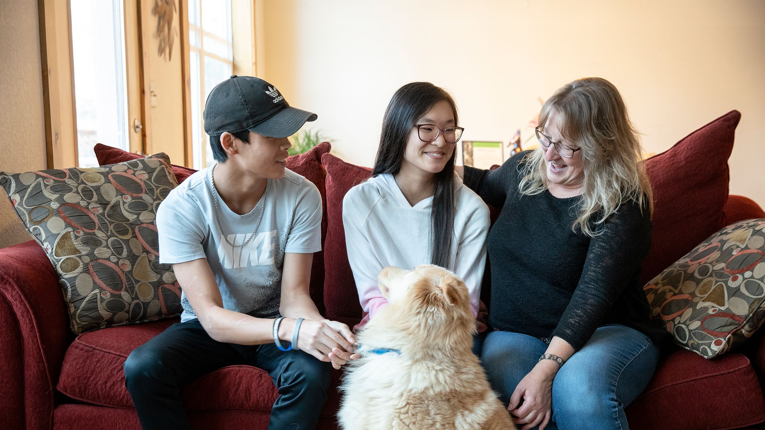 A woman and her two young adult children sit on their couch petting a golden retriever.