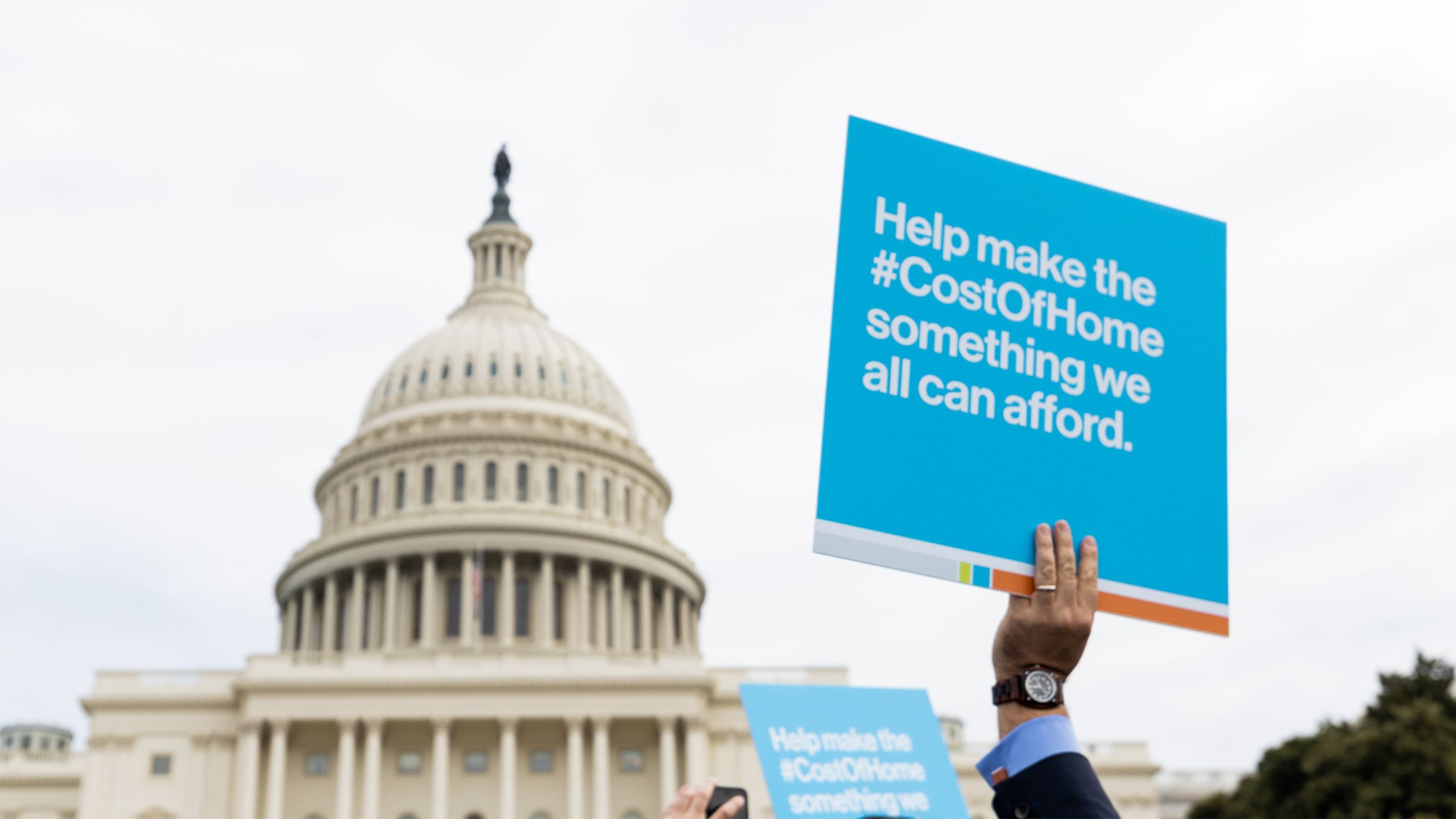 A hand holds up a protest sign reading "Help make the #CostOfHome something we all can afford" with the Capitol building in the background.