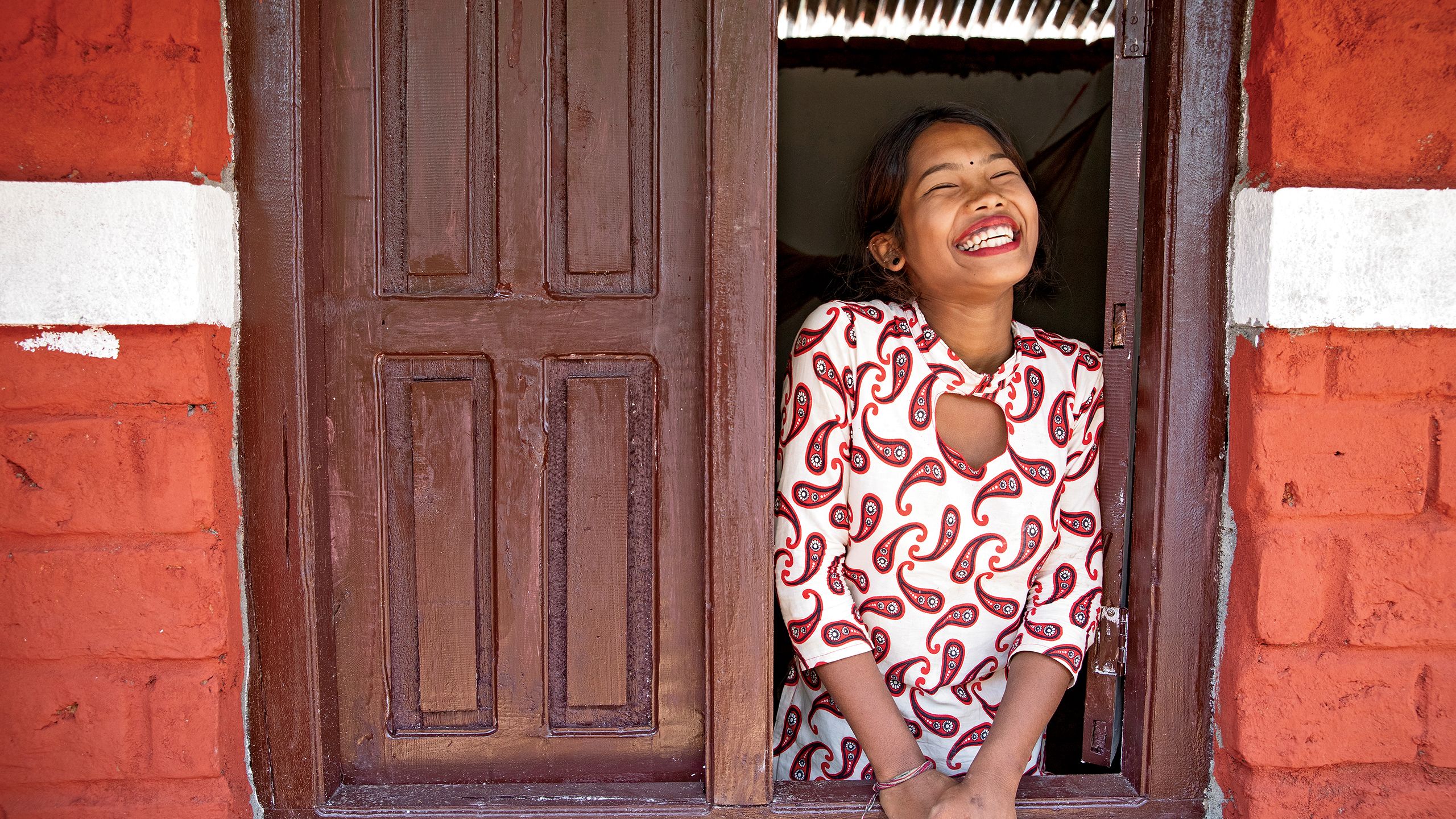 A young Nepalese woman beams as she leans out of the wooden window of her new, red-painted home.