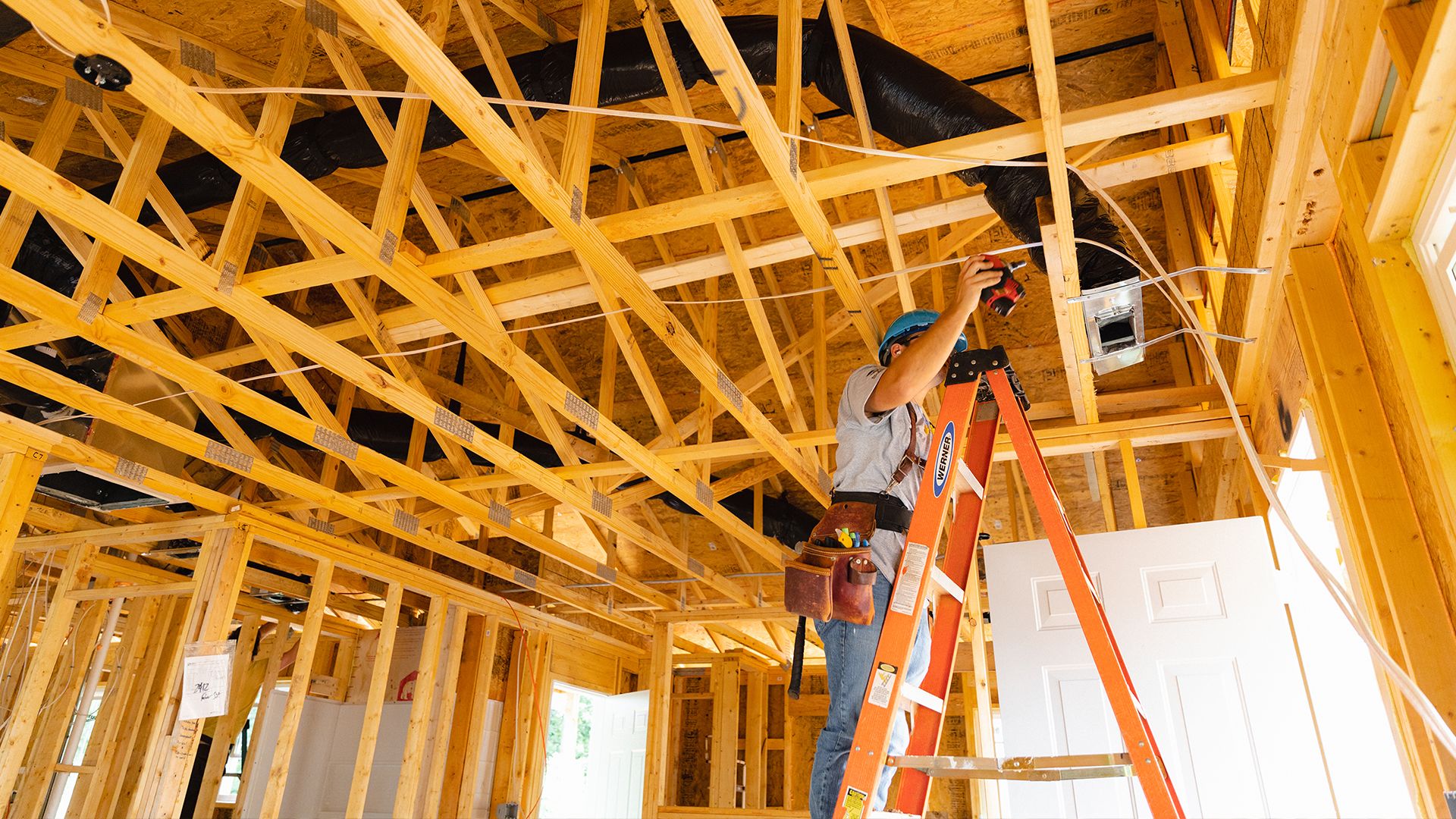 A volunteer stands on a ladder attaching wires through a fully framed ceiling.