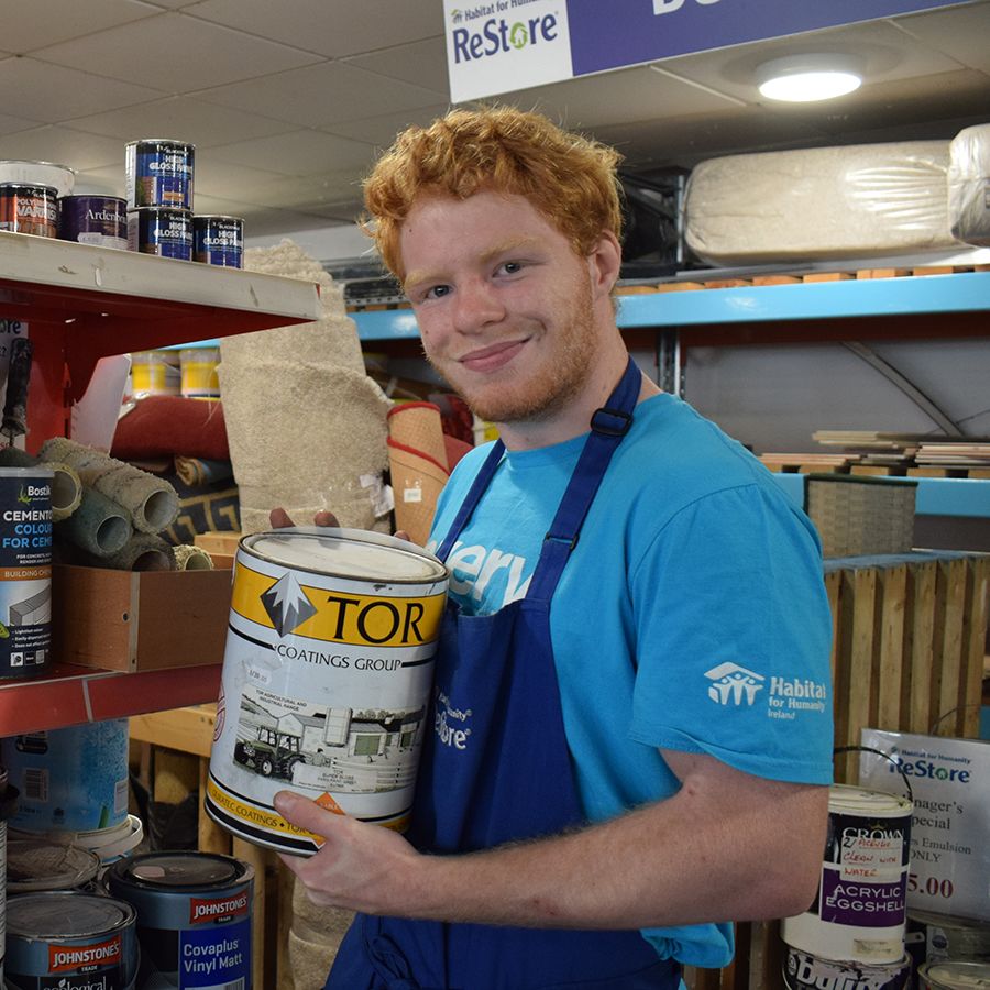 Jonny, a young red-haired man in a blue Habitat t-shirt and work apron, stocks paint on the shelves of the ReStore.
