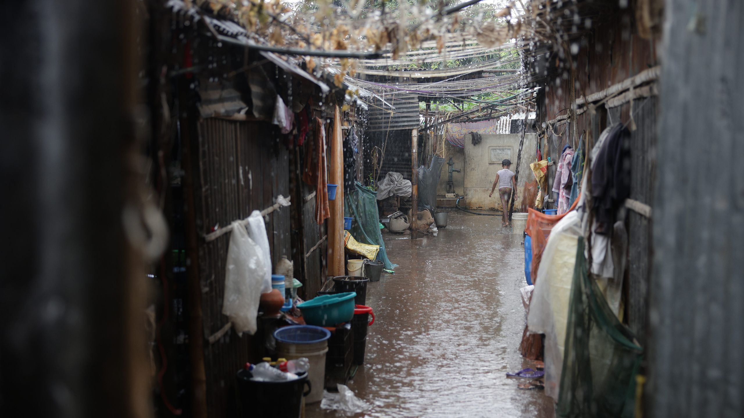 Rain drops from power lines draped across a flooding alley between houses. A young boy walks down the path without an umbrella.